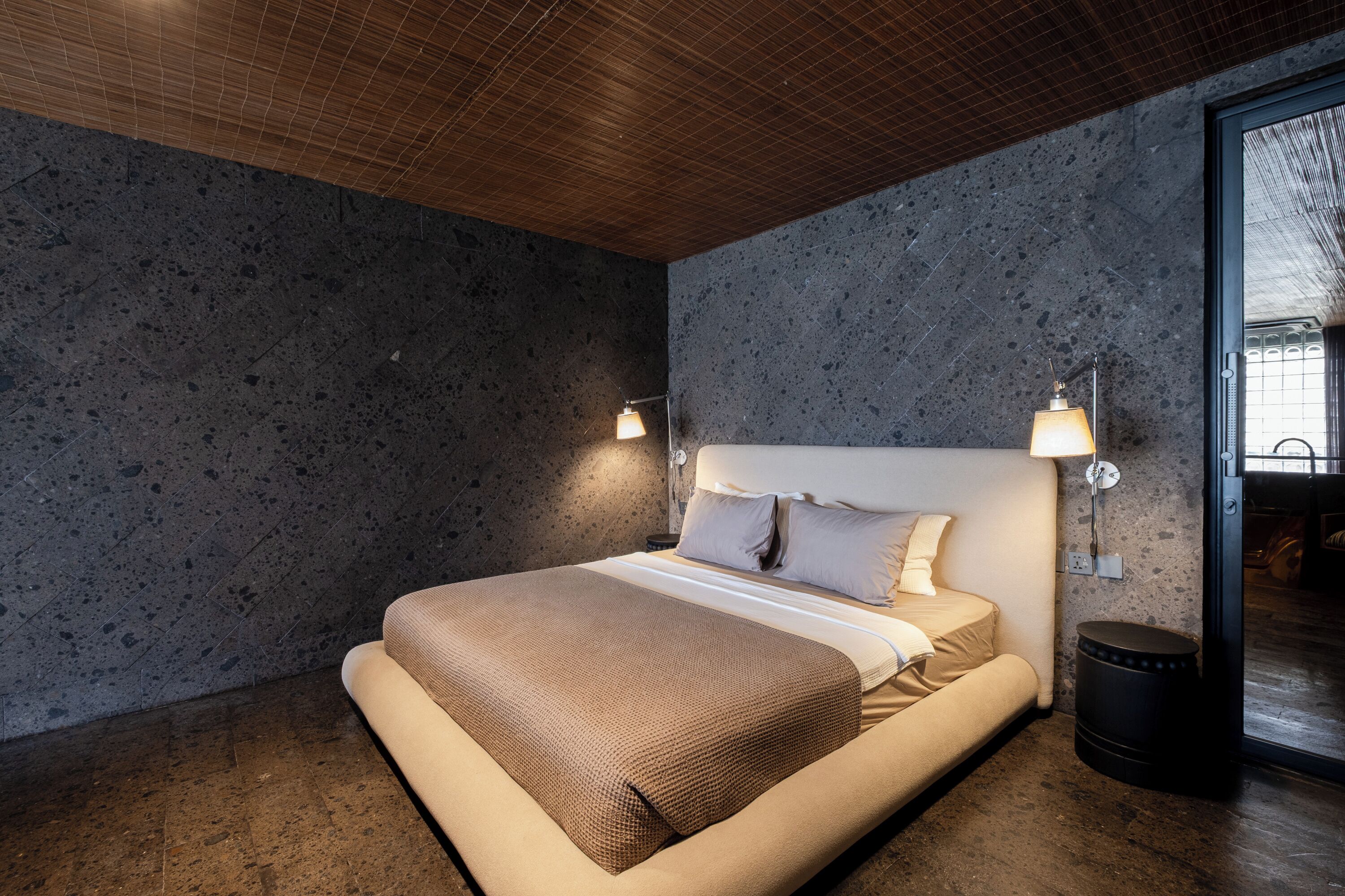 A modern bedroom with a large bed, two wall-mounted lamps, a textured wall, and a wooden ceiling.
