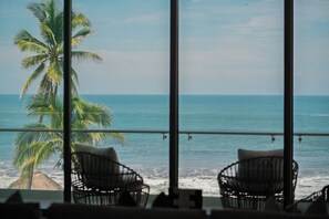 A balcony with two wicker chairs facing a palm tree and the ocean. - Every day begins with the Pacific just beyond your balcony. Sunset Surf is a spacious oceanfront residence in El Zonte featuring three bedrooms, panoramic ocean views, a resort-style pool, high-speed WiFi, and direct beach access for up to six guests (El Zonte)