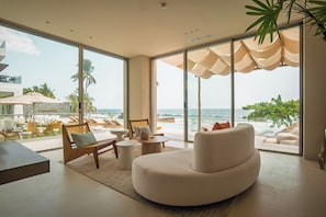 A modern living room with a large window, a white sofa, a wooden bench, and a small round table. - Every day begins with the Pacific just beyond your balcony. Sunset Surf is a spacious oceanfront residence in El Zonte featuring three bedrooms, panoramic ocean views, a resort-style pool, high-speed WiFi, and direct beach access for up to six guests (El Zonte)