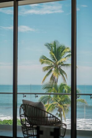 Ein Zimmer mit Meerblick und Palmen, ein Korbstuhl und eine Glasbarriere.