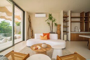 Ein modernes Wohnzimmer mit einer Couch, einem Couchtisch und Korbstühlen. Ein großes Fenster bietet einen Blick ins Freie.