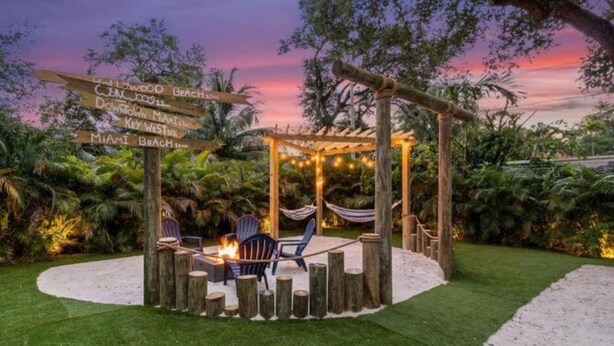 Un belvédère en bois doté d’un foyer et de chaises, entouré d’un jardin où un panneau indique les directions vers différents lieux.