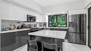 Une cuisine moderne dotée d’un grand îlot central, d’appareils électroménagers en acier inoxydable et offrant une vue sur la verdure environnante.