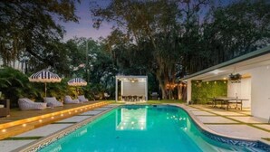 Une piscine avec des chaises longues et des parasols, un espace de repos couvert et un bâtiment doté d’un mur végétal et d’une fenêtre.