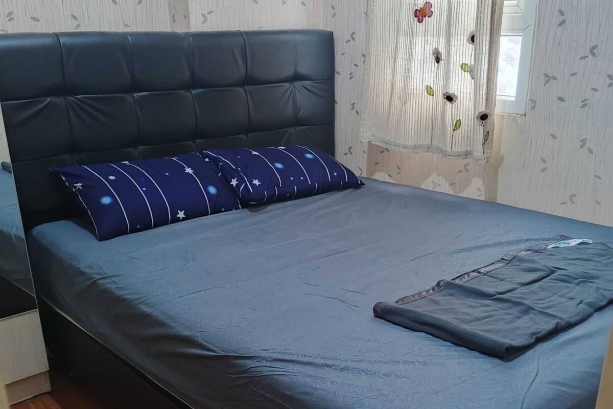 Een eenpersoonsbed met een donkerblauwe matras en passend kussen, een houten vloer en een raam met een bloemen gordijn.