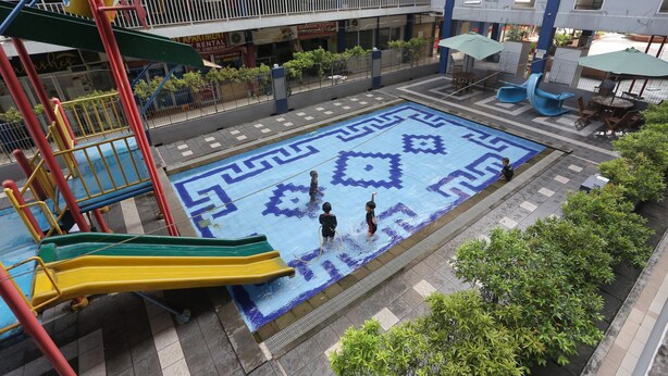 A playground with slides and a patterned play area, surrounded by a building with balconies and outdoor seating.