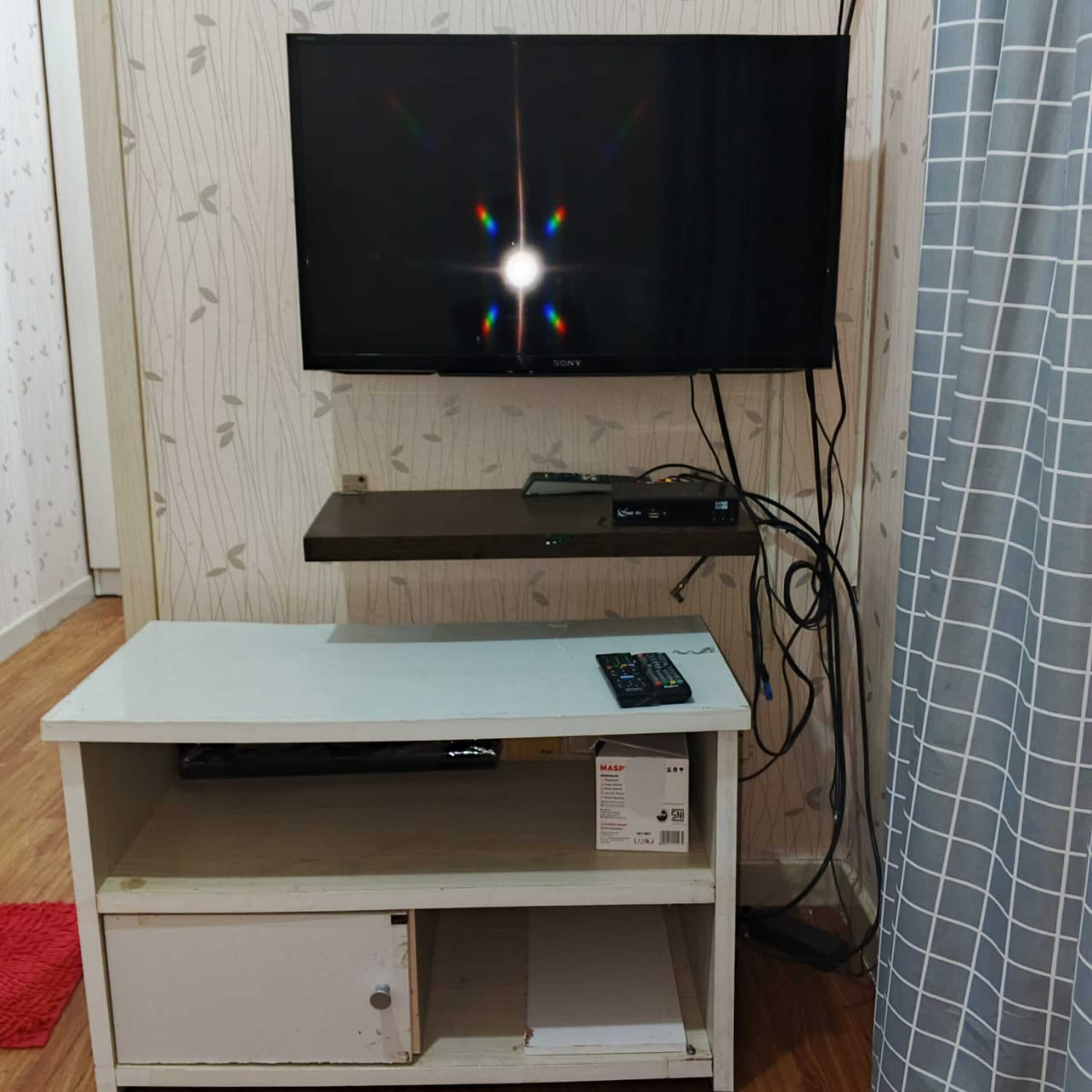 Un televisor montado en la pared con un estante debajo, un control remoto sobre el estante y una caja para organizar los cables.