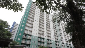 A multi-story residential building with balconies, a green facade, and a surrounding area with trees and a covered walkway.