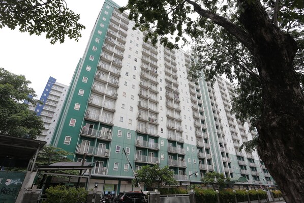 A multi-story residential building with balconies, a green facade, and a surrounding area with trees and a covered walkway.