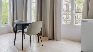 Une pièce avec une table et une chaise, une fenêtre avec des rideaux et une vue sur les bâtiments à l’extérieur.