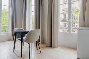 A room with a table and chair, a window with curtains, and a view of buildings outside. - Blueground | Le Marais, washer, nr Pompidou (Paris)