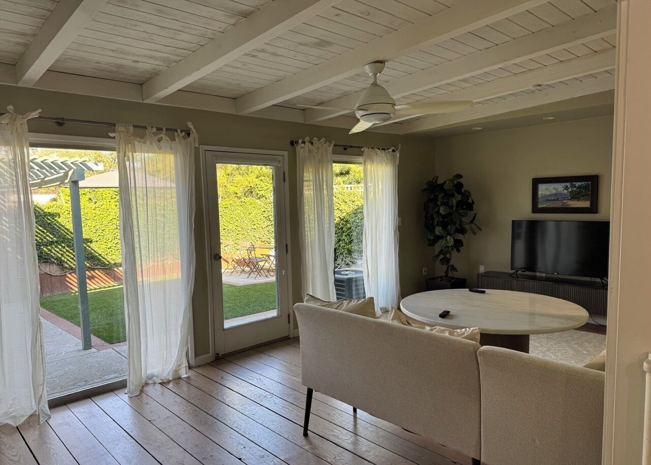 A room with a wooden floor, a round table, a sofa, a ceiling fan, and a TV.