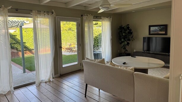 Una habitación con piso de madera, una mesa redonda, un sofá, un ventilador de techo y un televisor.