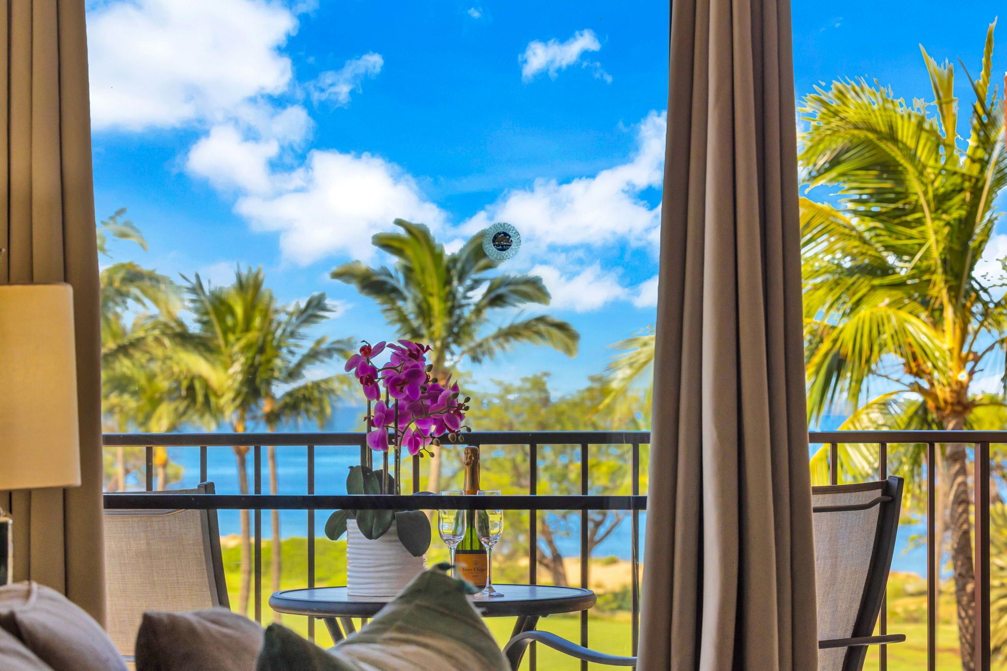 A balcony with a view of palm trees and the ocean, a table with a bottle and glasses, and a chair.