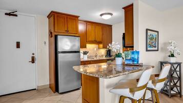 Una cocina moderna con armarios de madera, refrigerador de acero inoxidable, encimera de granito y una barra con sillas blancas.