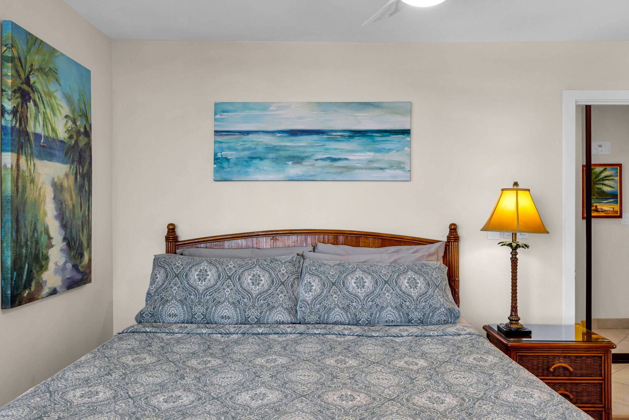Un dormitorio con una cama, un cuadro de una playa, una lámpara y una mesita de noche.