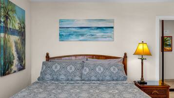 Un dormitorio con una cama, un cuadro de una playa, una lámpara y una mesita de noche.