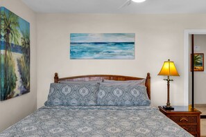 Un dormitorio con una cama, un cuadro de una playa, una lámpara y una mesita de noche.