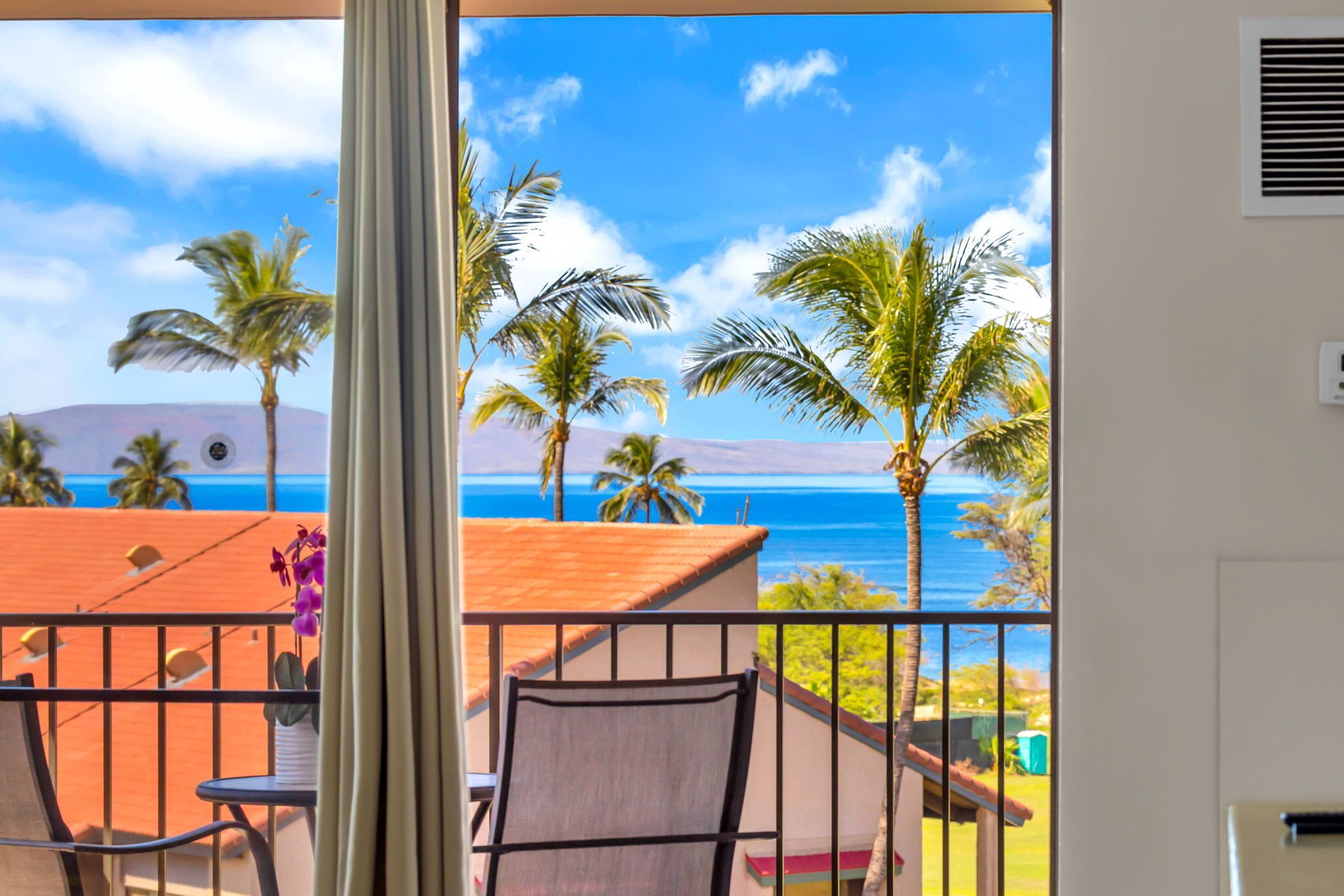 A balcony with a view of the ocean, palm trees, and a chair.