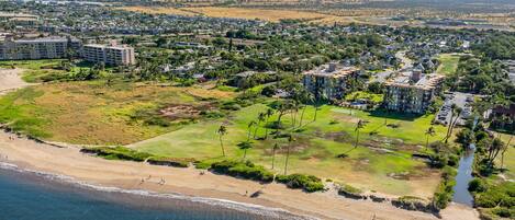 An aerial view of a coastal residential area with beachfront properties, a golf course, and a dense urban backdrop.