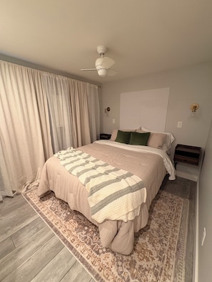 Ein Schlafzimmer mit einem Bett, Deckenventilator, Nachttisch, Wandlampe und einem Fenster mit Vorhängen.