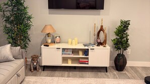 En stue med sofa, fjernsyn ophængt på væggen, et hvidt konsolbord med bøger og stearinlys, samt to krukkeplanter.