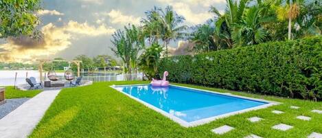 A rectangular swimming pool with a pink flamingo float, surrounded by well-manicured grass and palm trees.