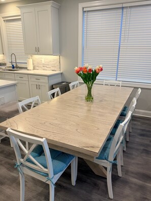 A dining area with a wooden table, white chairs with blue cushions, a vase of tulips, and a window with blinds.