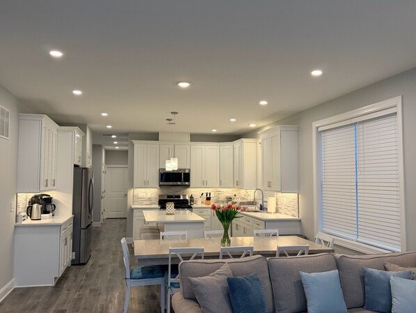 A modern kitchen with white cabinets, a dining area with a wooden table, and a gray sofa with blue cushions.