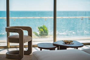 A modern room with a view of the ocean, a cushioned chair, a small wooden table, and a white sofa. - Welcome to Sun & Salt, an oceanfront condominium in El Zonte's Surf City featuring two bedrooms, two and a half bathrooms, a resort-style pool, and space for up to four guests. (El Zonte)