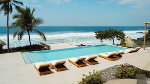 A pool with lounge chairs, palm trees, and a view of the ocean. - Welcome to Sun & Salt, an oceanfront condominium in El Zonte's Surf City featuring two bedrooms, two and a half bathrooms, a resort-style pool, and space for up to four guests. (El Zonte)