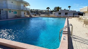 En swimmingpool med en metalstige, omgivet af liggestole og en bygning med altaner.