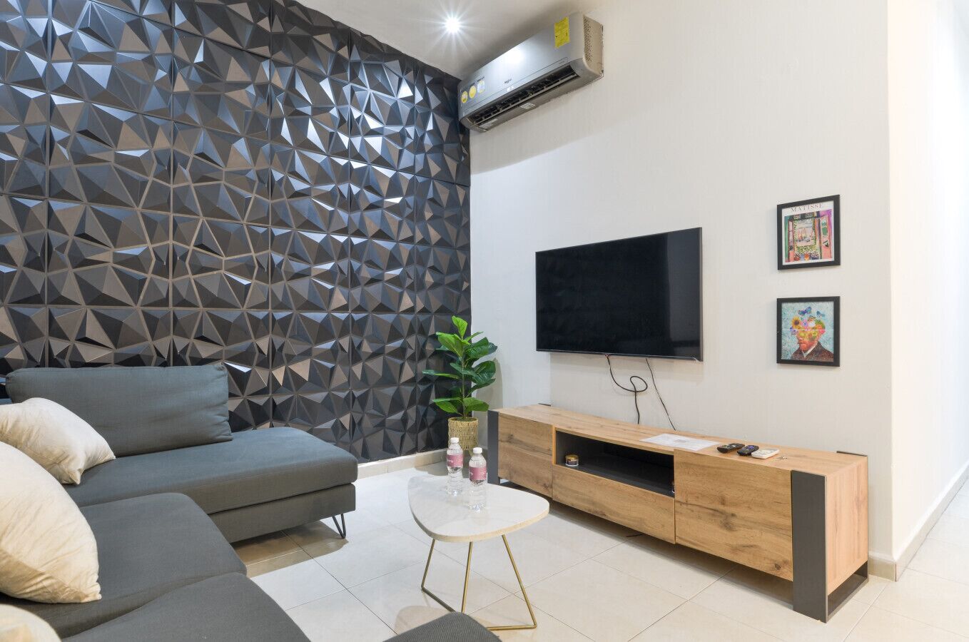A modern living room with a geometric-patterned wall, a grey sofa, a small white coffee table, a wooden TV stand, and a flat-screen TV.