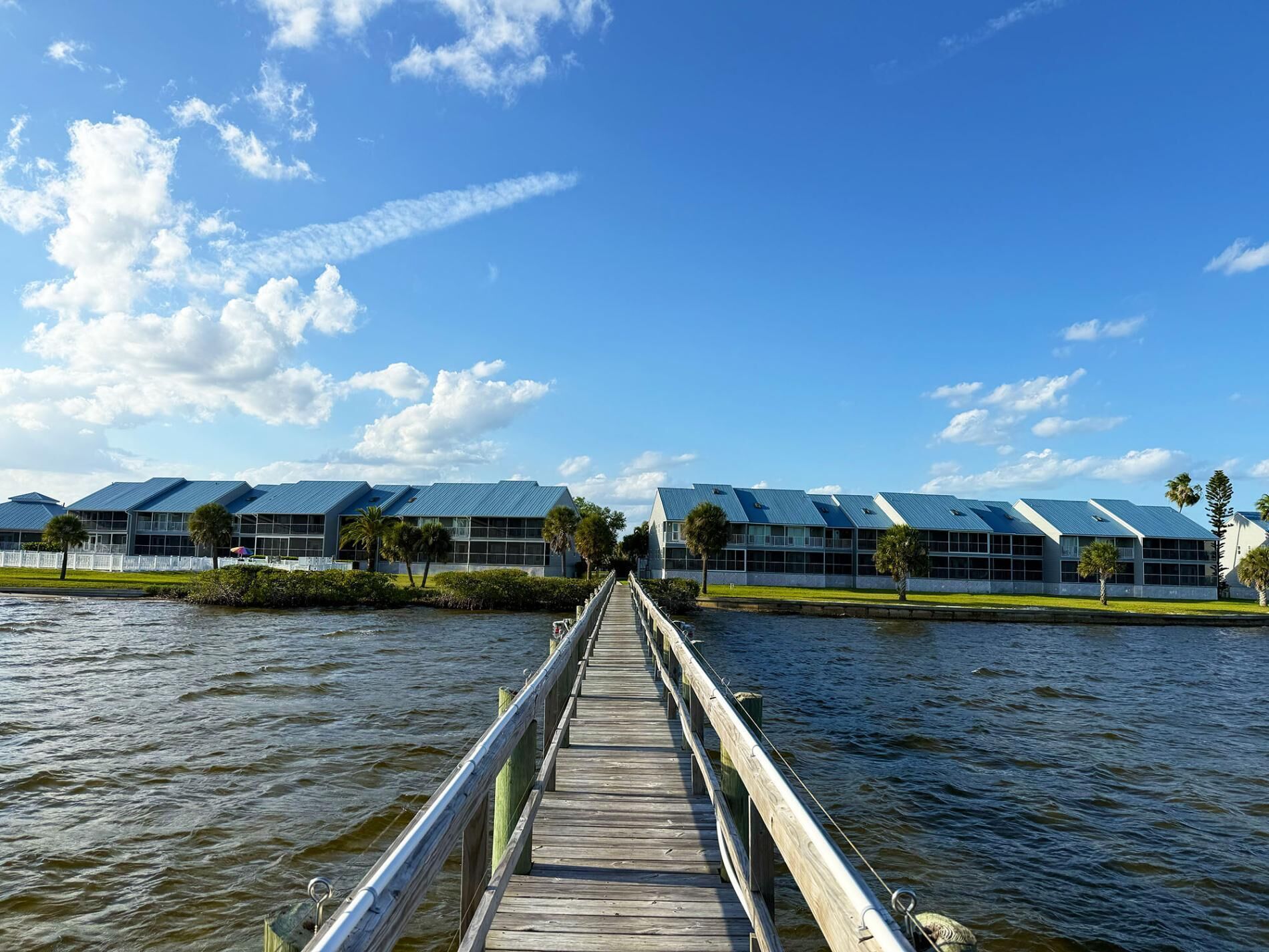 A wooden pier leading to a row of modern buildings with blue roofs, overlooking a body of water.