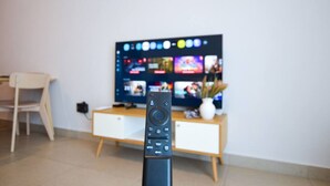 Een afstandsbediening in een kamer met een televisietoestel dat meerdere zenders toont, een witte stoel en een houten kast.