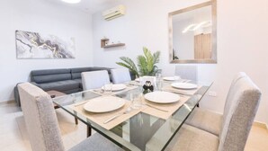 A modern dining area with a glass table, grey upholstered chairs, a grey sofa, a wall-mounted air conditioner, and a framed mirror.