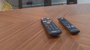 A television mounted on a wall displaying a streaming service interface. Two remote controls are on a wooden table in front of the TV.