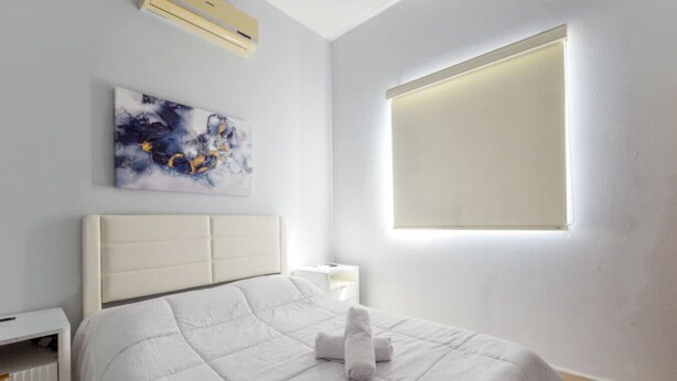 A bedroom with a bed, a nightstand, an air conditioner, a window, and a painting on the wall.
