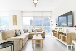 Ein modernes Wohnzimmer mit einem Ecksofa, einem Couchtisch, einem Fernseher und einem großen Fenster mit Blick auf die Stadt.