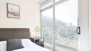 Une chambre avec une grande fenêtre, un lit, une table de chevet avec une lampe et un tableau encadré au mur.
