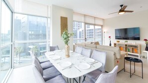 Un espace repas moderne avec une table dressée pour quatre personnes, un canapé et un téléviseur, le tout dans une pièce lumineuse dotée de grandes fenêtres.
