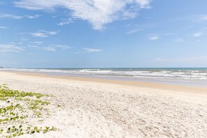 Ein Strand mit Sand und Meer, bewölkter Himmel und etwas Vegetation.