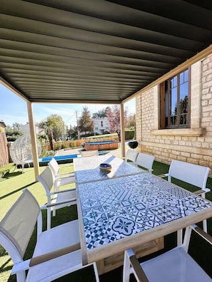 Eine Terrasse im Freien mit überdachtem Tisch und Stühlen, Blick auf einen Swimmingpool und ein Steinhaus im Hintergrund.