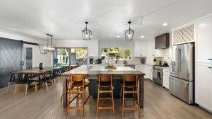 A modern kitchen with a central island, bar stools, a dining table, and a large refrigerator. - Tropical Villa by Glam Homes (Scottsdale)