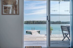 En vy genom en glasdörr mot en träbrygga som sträcker sig ut i en sjö, med en bänk och en krukväxt på bryggan.