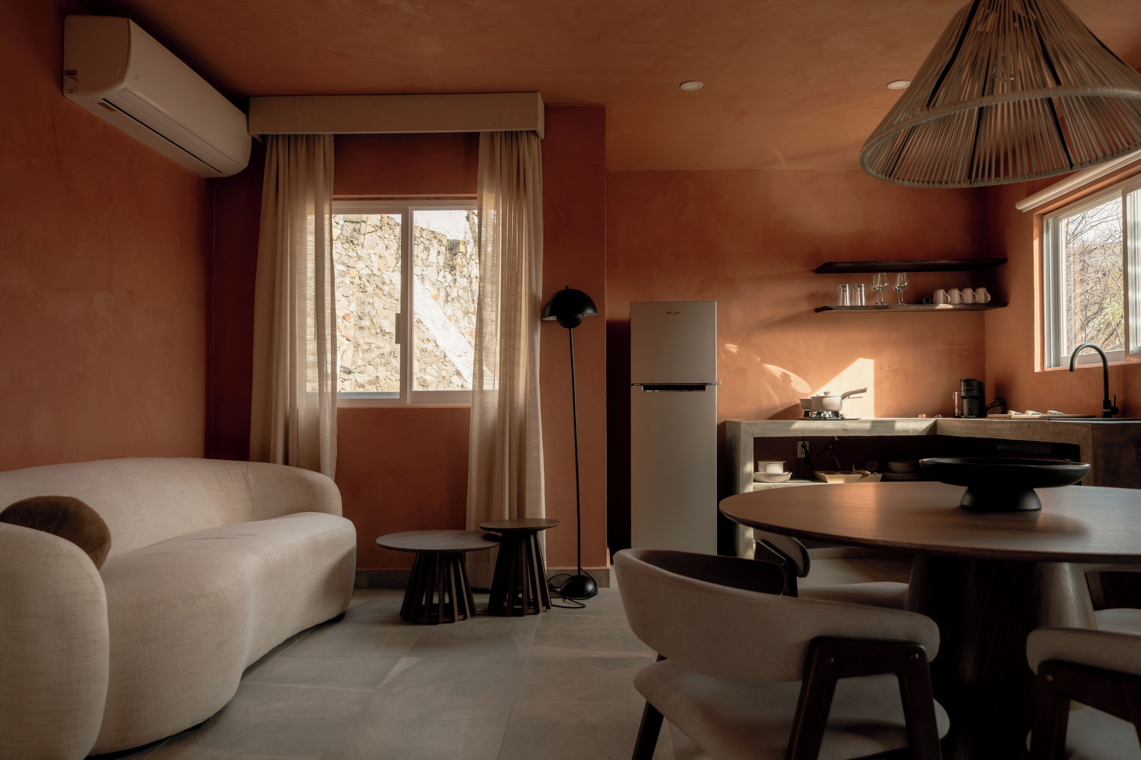 Un espacio habitable compacto con sofá, mesa de comedor, pequeña cocina y un amplio ventanal con cortinas.