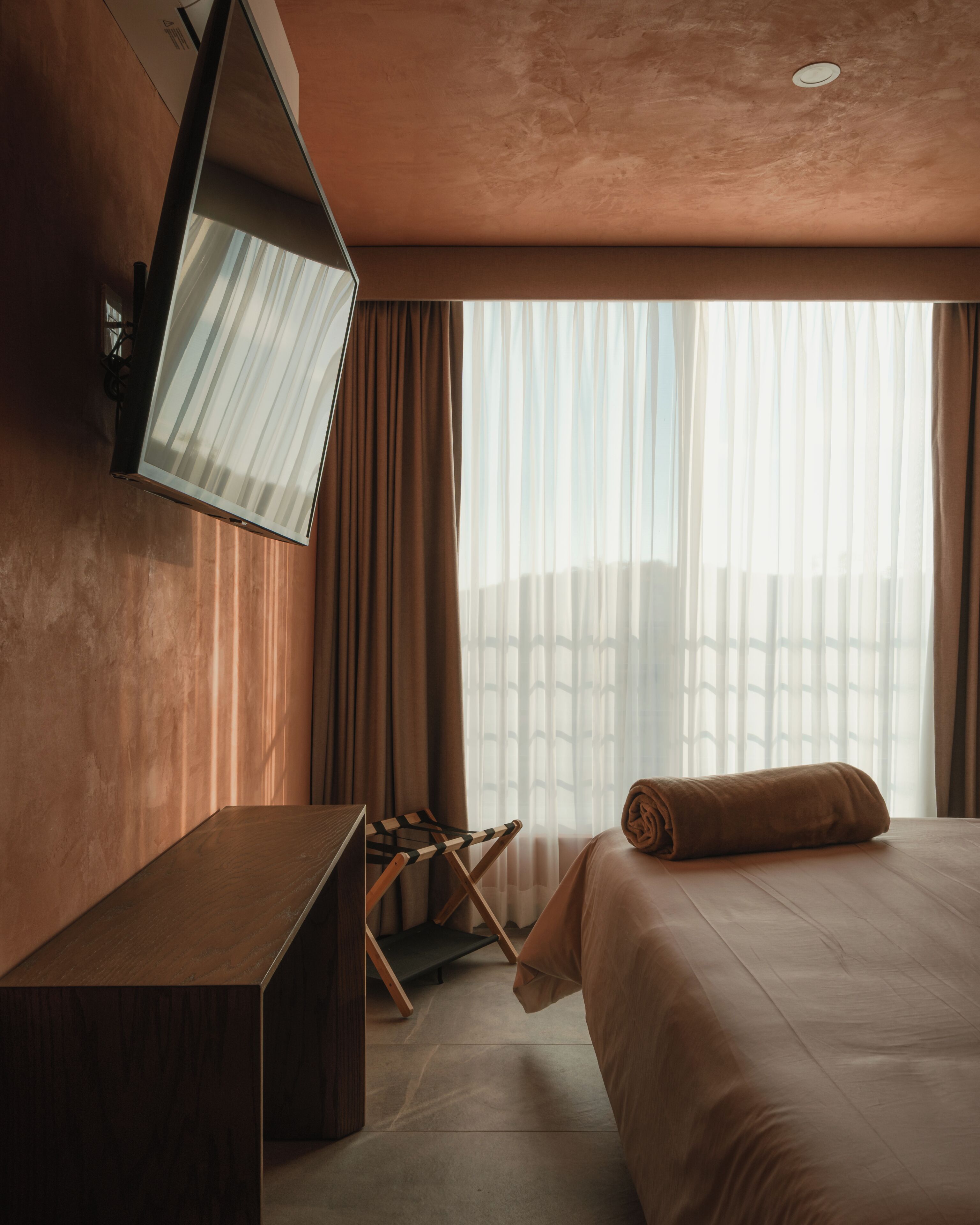 A hotel room with a bed, a desk, a chair, a television mounted on the wall, and a window with curtains.