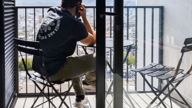 Une personne assise sur une chaise de balcon, en train de parler au téléphone, avec une vue sur la ville et des montagnes en arrière-plan.