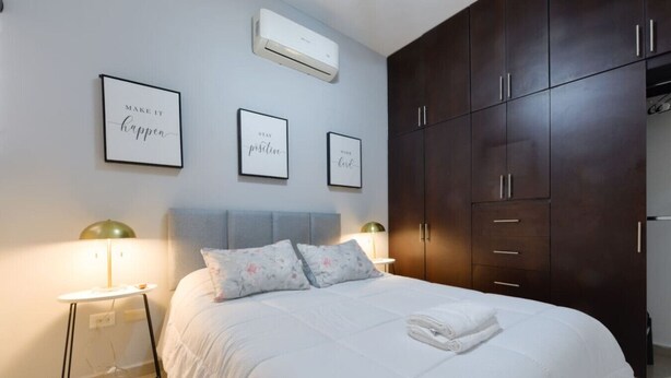 Quarto com cama, luminárias de cabeceira, guarda-roupa e arte motivacional na parede.