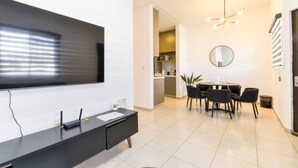 Uma sala de estar moderna com uma televisão de tela plana, uma área de jantar com mesa e cadeiras redondas, e uma cozinha visível por uma porta aberta.